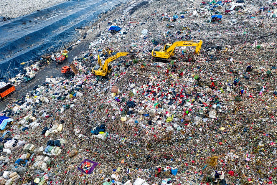 An expansive landfill site filled with layers of mixed waste, including plastic bags, fabric, and various household debris resting on a large, sloped area. Two yellow excavators with long arms and bucket attachments are actively engaged in compacting or removing rubbish, positioned towards the right and center of the image. The waste is scattered across the uneven terrain, creating mounds and valleys, with some areas covered by black plastic sheeting on the left side of the scene. In the background, faint figures of workers or machinery may be visible amidst the chaos of disposed materials. The environment appears outdoor, with natural lighting highlighting the textures and colors of the waste, such as bright reds, blues, and whites contrasted against more muted earth tones. This scene demonstrates a typical example of waste management through on-site bulk removal, aligning with independent rubbish clearance services like those provided by Rubbish Clearance Ilford, emphasizing the scale of waste disposal activities in urban or industrial settings.