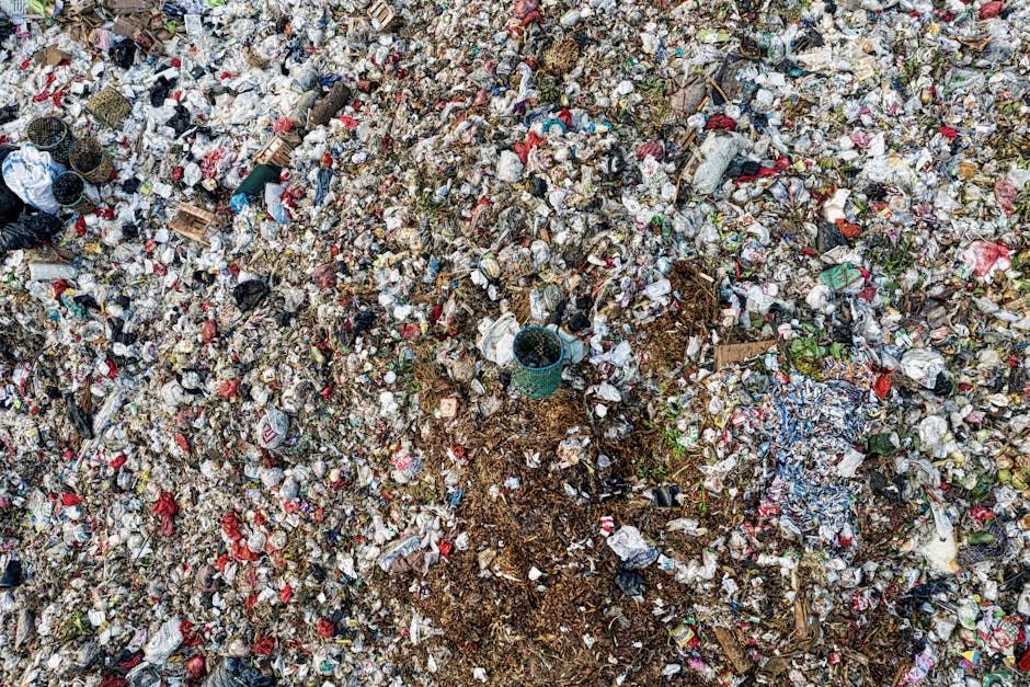 An expansive view of a large, unmanaged landfill site filled with a dense mixture of waste materials. The surface is covered with various types of rubbish, including plastic bottles, food packaging, paper, cardboard, and discarded miscellaneous items, creating a chaotic and cluttered landscape. Some plastic bottles are transparent or colored, with caps visible among other debris. Twisted paper and torn cardboard pieces are scattered throughout the heap, alongside bits of organic waste and soil. In the middle of the scene, a small, round, green plastic container partially buried among the rubbish, indicating improper disposal. The background reveals a broad, uneven terrain of waste extending to the horizon, with no visible boundaries or containment structures. The overall environment appears outdoors under natural lighting, contributing to a natural, unregulated waste accumulation site. Such scenes exemplify challenges in waste management and highlight the importance of proper rubbish removal services, such as those provided by Rubbish Clearance Ilford, to tackle alternative waste handling needs beyond typical council collection methods.