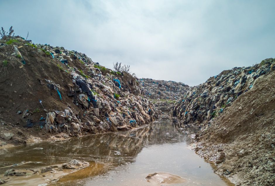 An outdoor scene depicting a narrow, uneven trench filled with stagnant, muddy water. The sides of the trench are composed of piled-up soil and debris, including various pieces of plastic waste such as bags and fragments in shades of blue, black, and transparent. The soil appears dry and compacted with rough, rocky textures, contrasting with the slick, wet surface of the water within the trench. Sparse small plants and weeds are visible on the upper edges of the soil mounds, indicating a minimally vegetated environment. Overhead, the cloudy sky suggests overcast weather, casting diffuse lighting over the scene. The environment appears to be part of a neglected or unmanaged area of land, illustrating the impact of improper waste disposal and the importance of professional rubbish removal services like those offered by Rubbish Clearance Ilford, which may intervene in such contexts to facilitate cleanup and proper waste handling.