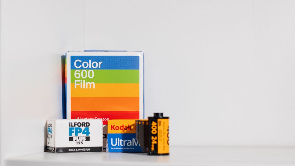 A collection of photographic supplies arranged on a plain white surface against a white background, including a stack of three boxes: a large box of Kodak Ultra Max 400 35mm film with orange, black, and yellow branding, a smaller box of ILFORD FP4 Plus black and white film with predominantly white packaging and blue and black accents, and a larger box of Color 600 Film featuring bold blue, green, orange, and red stripes. In front of the boxes, there are three small rolls of photographic film, with visible black and orange protective packaging. The arrangement appears organized, with the larger boxes positioned at the back and the smaller rolls in front, capturing a close-up view suitable for an article about alternative waste handling or private collection of photographic materials, in alignment with rubbish removal services like those offered by Rubbish Clearance Ilford. The scene is evenly lit, highlighting the textures of the cardboard boxes and the glossy film rolls, and suggesting a tidy, indoor environment typical for storage or collection of photographic equipment and materials.