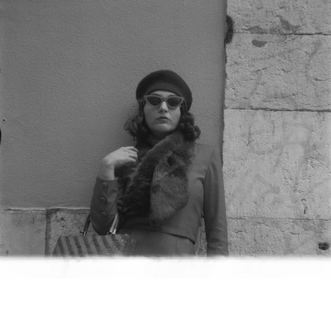 A black-and-white photograph featuring a woman standing outdoors against a textured stone and plaster wall. She is dressed in vintage-style clothing, including a tailored coat with buttons, a fur stole draped over her shoulders, and a dark beret on her head. She wears large, rounded sunglasses and has shoulder-length wavy hair. She is holding her left hand near her neck, with a subtle expression on her face. In front of her, at the bottom edge of the image, part of what appears to be a horizontal surface is visible, possibly a ledge or table. The setting suggests an urban environment, with the stone wall providing a textured background. The overall scene has a stark, classic feel that could be associated with urban fashion or historical street photography, relevant to discussions of city environments, private appearances, or independent personal styles, which are sometimes encountered in contexts related to local waste and rubbish removal services like those operated by Rubbish Clearance Ilford, especially in outdoor, urban areas.