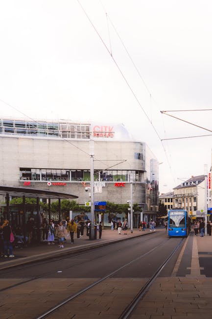 A busy urban street scene featuring a modern multi-storey building with large glass windows and concrete walls in the background. In the foreground, railway tracks run parallel to a paved sidewalk where a group of pedestrians is waiting under a bus shelter. The shelter has a curved roof supported by metal poles, with seating and a small number of people standing nearby. To the right, a blue tram is approaching along the tracks, framed by overhead electric wires extending across the scene. The surrounding environment includes adjacent buildings with traditional facades, signage, and trees lining the sidewalk. The lighting suggests an overcast day with diffused natural light, creating a neutral and professional atmosphere consistent with a city centre location. The scene subtly hints at the context of public transportation although it is not a direct visual of waste management or rubbish removal services, but could relate to alternative urban transit options including private collection or city infrastructure supporting urban logistics.