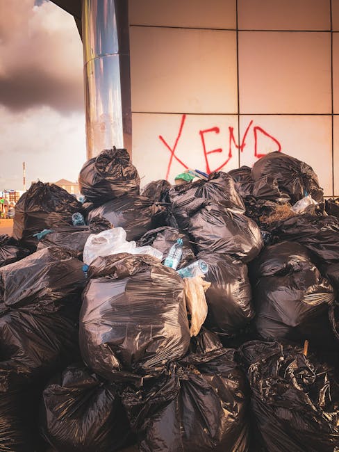 A large accumulation of black waste disposal plastic bags filled with rubbish positioned against a tiled wall of modern architecture, with some of the bags partially torn or crumpled, revealing mixed contents. Among the bags are visible discarded plastic water bottles, some crushed and scattered around the base. The background features graffiti painted in red, with the word 'XEND' visible on the wall. The scene is outdoors on a paved surface, with an overcast sky casting diffused light over the area. This image illustrates a pile of waste awaiting collection, relevant to private rubbish removal services offered by companies like Rubbish Clearance Ilford. The presence of such waste on streets or urban areas often highlights the necessity for specialist waste clearance, especially when local authority collection services are not involved, emphasizing the importance of professional rubbish disposal and on-site clearance options.