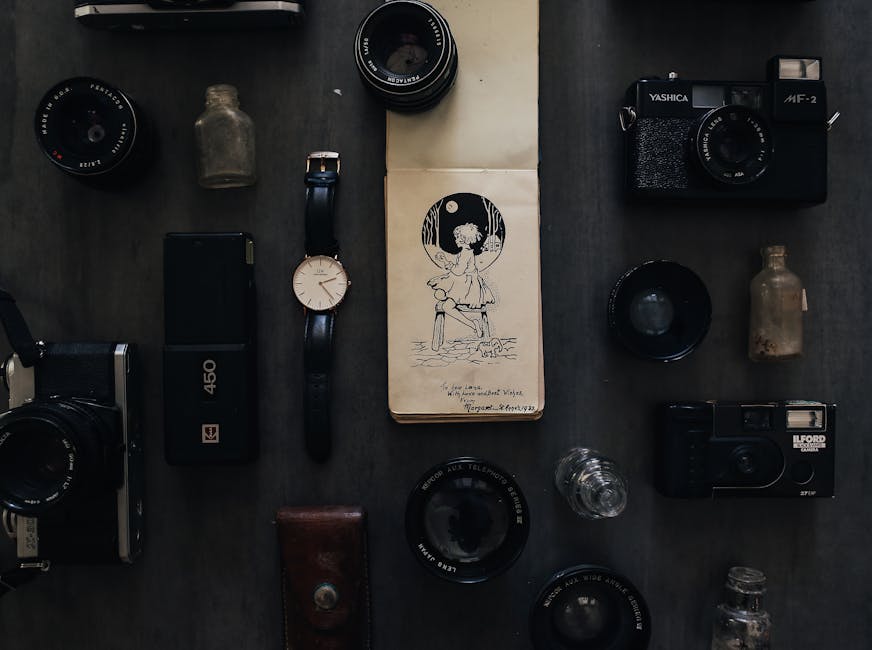 An overhead view of a collection of vintage cameras, lenses, and photography accessories arranged on a dark, textured surface, with some items placed on top of a sealed cardboard package featuring a black-and-white illustration of a girl sitting on a dock by a body of water under a full moon. The cameras include a rangefinder-style model with silver and black finishes, a compact point-and-shoot camera, and an older film camera with a leather strap. Several camera lenses of varying sizes and focal lengths are also visible, some with protective caps. There are two small glass bottles, one appearing weathered and slightly discolored, positioned near the camera equipment. A wristwatch with a white face and black leather strap, along with a portable power bank, are placed among the gear. The scene suggests a curated display of photography equipment, which could be related to private collections or alternative equipment storage, in a setting that might be used for photography or equipment maintenance, reflecting a focus on preserving or handling vintage and modern camera gear.