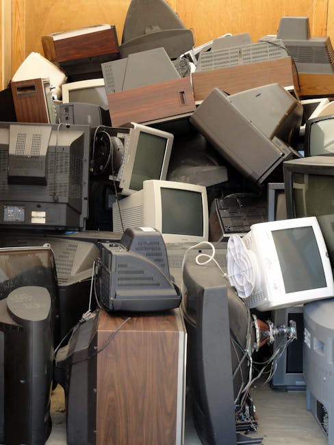 A large pile of discarded electronic waste, primarily consisting of vintage computer monitors and CRT screens with bulky, box-like shapes and dark grey or beige plastic casings. Some monitors are stacked on top of each other or leaning against a wooden wall in the background, creating a dense, cluttered scene. In the foreground, several old computer towers and peripherals, such as keyboards and cords, are visible among the monitors, with various textures including smooth plastic and ribbed ventilation panels. The environment appears to be an indoor storage space or waste collection area, with natural light illuminating the scene and emphasizing the mix of materials and muted colour tones. The image illustrates the type of waste that may require professional rubbish clearance services, such as those offered by Rubbish Clearance Ilford, to facilitate proper disposal or recycling of obsolete electronics in line with waste management regulations.