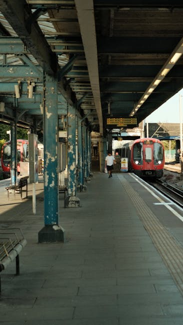 The image depicts a railway station platform with an overhanging roof supported by blue-painted metal pillars showing signs of paint chipping and weathering. The platform surface is paved with concrete slabs, featuring tactile paving strips along the edge for safety. A red passenger train is stationed on the tracks to the right, with its doors closed. A person dressed in white clothing is walking along the platform, moving away from the camera towards the train, while another underground train is visible further along the tracks on the right side. The surrounding environment includes a partial view of a station sign and some lighting fixtures attached to the roof structure. The scene is illuminated by daylight, casting soft shadows, and the station appears clean and well-maintained. This setting, typical of an urban railway station, relates to alternative methods of transporting waste or debris that require removal, aligning with services like those offered by Rubbish Clearance Ilford for private rubbish disposal and on-site clearance solutions, although no specific waste objects are visible in this image.