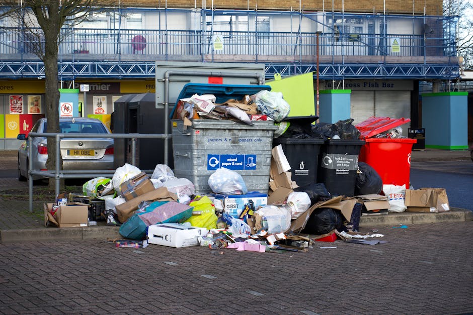 A collection of overflowing rubbish bins and scattered waste is located on a paved urban street beyond a low metal barrier. The waste includes various black and red plastic bin bags, flattened cardboard boxes, paper and plastic packaging, and loose items like cardboard tubes and small household objects. The central bin, labeled for mixed paper and card, is partially open with crumpled paper, documents, and flattened cardboard spilling onto the ground in front of it. To the right, a green recycling bin and a black bin are also visible, with additional discarded materials piled around their bases. Behind the waste area, there are commercial storefronts with yellow and white signage, and a building under construction covered in blue scaffolding and protective netting. The scene is illuminated by natural daylight, highlighting the textures of the trash and the surrounding urban environment, demonstrating typical private waste accumulation that might be addressed through services like those offered by Rubbish Clearance Ilford for alternative waste handling and rubbish removal.