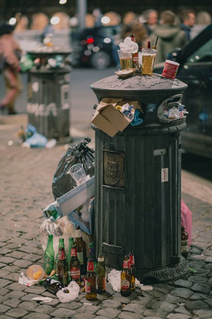 A black, vintage-style public rubbish bin made of metal with a textured, slightly weathered finish is situated on a cobbled sidewalk in an urban area. The top of the bin is covered with various discarded items, including used paper cups with lids, some containing remaining drinks, and a crumpled takeaway food container. Several empty glass bottles, predominantly brown and green, lie stacked at the base of the bin, with some tipped over and others partially crushed. Loose litter such as used napkins, food wrappers, and torn paper are scattered around the bottom and on the ground nearby. In the background, blurred figures of pedestrians walk past another bin and a black car is partially visible parked on the street. The scene suggests a busy outdoor setting where waste has accumulated due to high foot traffic, highlighting the importance of effective rubbish removal and private waste handling services like those offered by Rubbish Clearance Ilford.