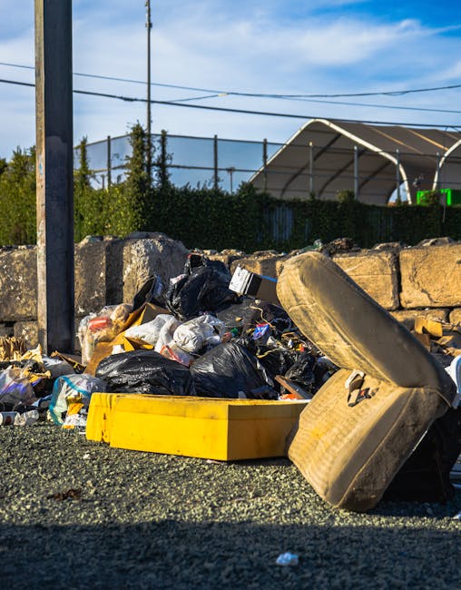 A pile of mixed waste materials including black plastic garbage bags, a yellow plastic container, and a discarded car seat with beige upholstery and dark support structure, all set on a gravel surface. The rubbish is situated against a weathered stone wall, with a utility pole and overhead power lines visible to the left. In the background, there is a tall hedge, some trees, and a large covered sports or storage structure with a curved metallic roof, set against a partly cloudy sky. The scene appears to be an outdoor area designated for rubbish accumulation, possibly indicating a private or alternative waste collection site, as managed by Rubbish Clearance Ilford, aligning with local rubbish removal services and on-site clearance practices.