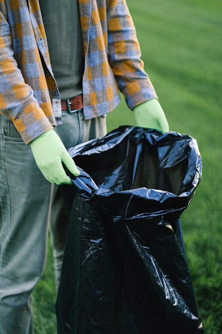 Close-up of a person standing outdoors on a grassy area, wearing a yellow and grey checkered shirt over a green top, with a brown belt and grey trousers. The individual is holding a large black garbage bag open, lined with a shiny black plastic interior, and is wearing light green disposable gloves. The bag appears to be partially filled or prepared for rubbish collection, with the surrounding environment suggesting a garden or park setting. The scene emphasizes responsible waste handling and collection, indicative of private rubbish clearance services offered by Rubbish Clearance Ilford, which may include on-site waste disposal or alternative rubbish removal options outside traditional council collections. Natural daylight illuminates the scene, highlighting the textures of the clothing and the bag, while the background shows a blurred view of the green lawn, emphasizing a focus on waste management activity in an outdoor, environmentally conscious context.