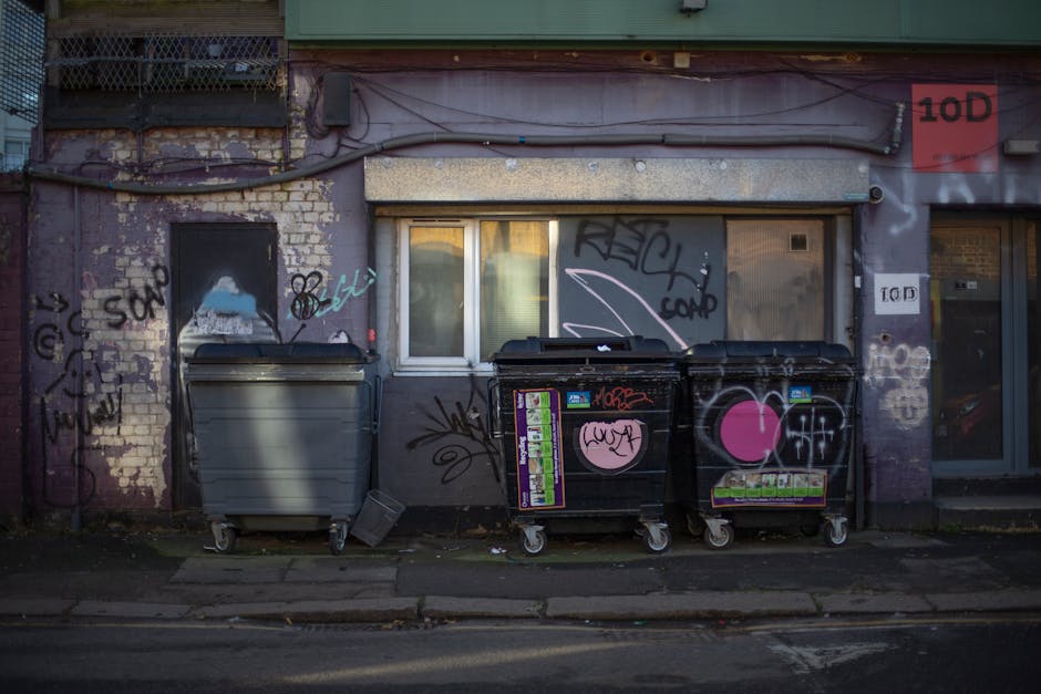 The image depicts a section of an urban building exterior with a façade displaying weathered brickwork and a faded purple wall covered in various graffiti tags and spray-painted symbols. In front of the wall, there are three large wheeled waste bins positioned along the sidewalk. The leftmost bin is grey with a closed lid, while the other two are black with bright pink heart-shaped stickers and additional colorful stickers, all with closed lids. Nearby, a small, grey, rectangular object lies on the ground, leaning against the grey bin. The building features a small, rectangular window with a white frame, partially obscured by the bins, and a rolled-up metallic shutter above it, suggesting an entryway. To the right, a glass door with a dark frame is visible, with the building signage visible on small panels. The environment implies a typical urban alleyway or street corner landscape, which may relate to independent waste collection or private rubbish disposal services carried out by companies like Rubbish Clearance Ilford, evident through the presence of the bins designated for rubbish collection.