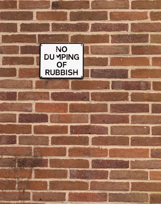 A rectangular metal sign mounted on a red brick wall with horizontal mortar joints, displaying the message 'NO DUMPING OF RUBBISH' in black uppercase letters on a white background. The sign is positioned slightly above the center of the image, with the brick wall extending evenly across the frame. The wall's surface shows a consistent pattern of reddish-brown bricks with some variation in tone, and the mortar appears light grey. The lighting is natural, casting minimal shadows, and the overall scene suggests an area designated to discourage illegal rubbish dumping, aligning with practices for private waste management and rubbish clearance services such as those offered by Rubbish Clearance Ilford.