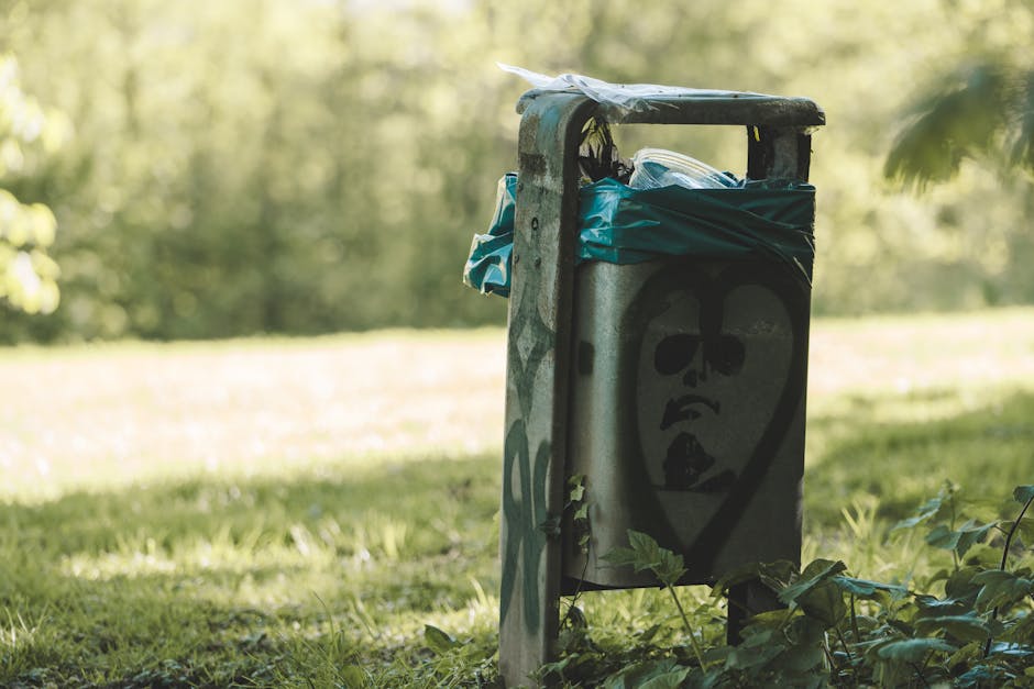 A metallic public litter bin situated on a grassy area outdoors, with a spray-painted black face featuring large oval eyes, a small nose, and an open mouth in a graffiti style. The bin has a weathered, slightly rusted finish and is partially covered with a blue plastic bag draped over the top for containment. Surrounding the bin, there are green leaves and small plants at its base, and the background shows a blurred natural environment with trees and sunlight filtering through the foliage. The overall scene suggests an urban park or communal outdoor space where informal waste disposal occurs, and the presence of graffiti indicates independent collection or non-official waste handling typically associated with rubbish removal services like those offered by Rubbish Clearance Ilford.